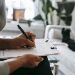 Unrecognizable ethnic female therapist taking notes on clipboard while filling out form during psychological appointment with anonymous client lying on blurred background